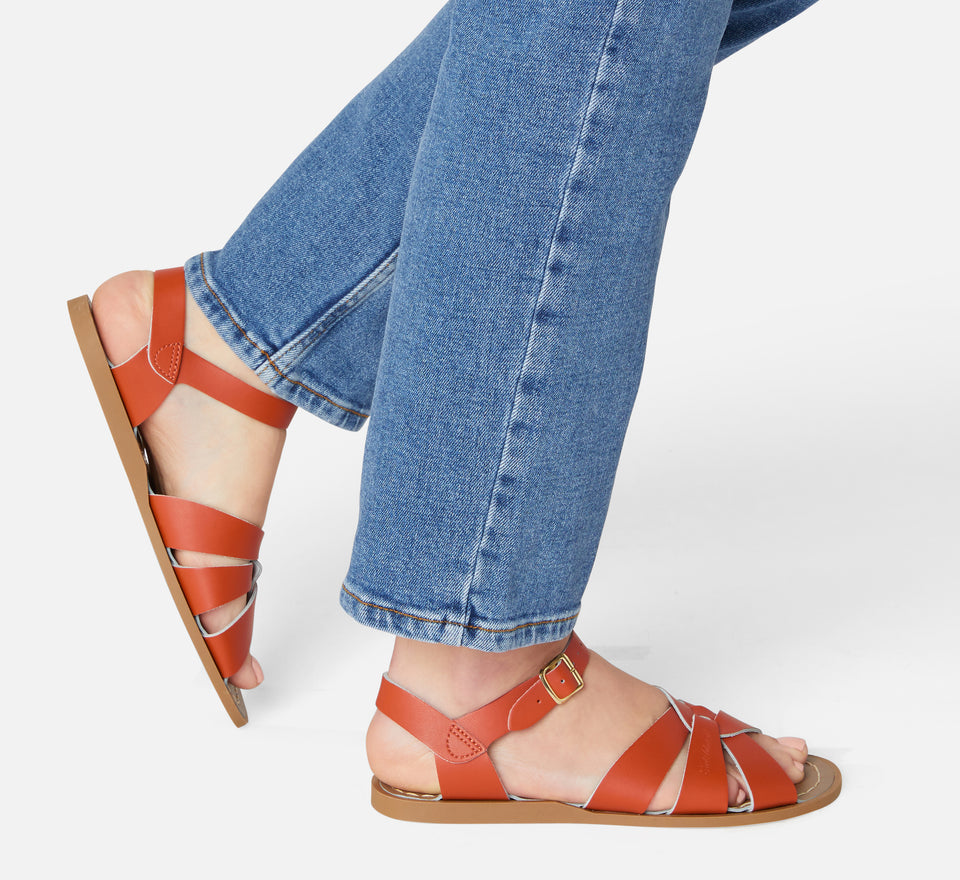 An original girls sandal by Salt Water Sandals in paprika with a single buckle fastening around the ankle. Open Toe and Sling-back with woven detail on the front. Lifestyle of walking view.