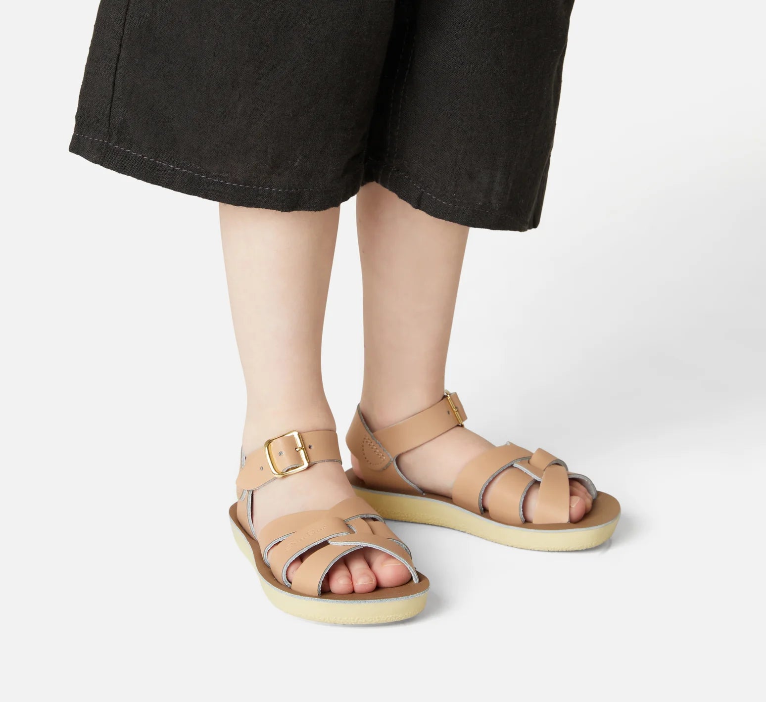 A unisex sandal by Saltwater, style swimmer in Latte with gold buckle strap. Image of child modelling the sandals