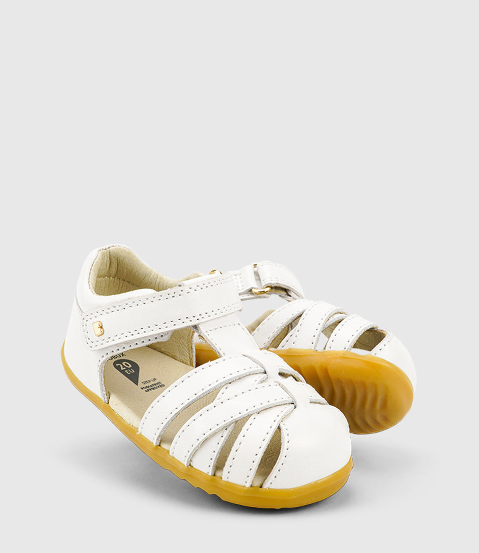 A unisex sandal by Bobux, style Cross Jump in white with velcro strap and light brown sole. Tilted view of pair