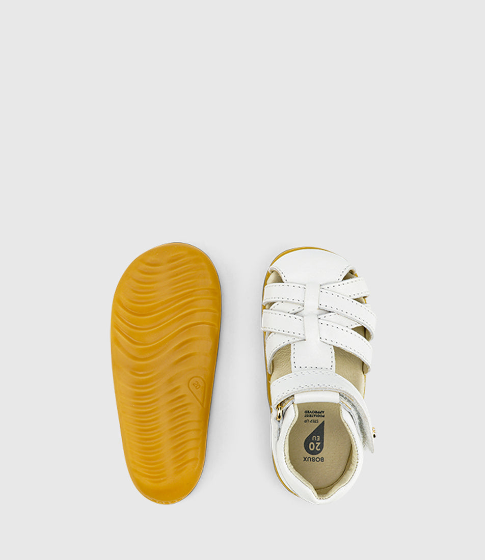 A unisex sandal by Bobux, style Cross Jump in white with velcro strap and light brown sole. View of pair with one sole showing