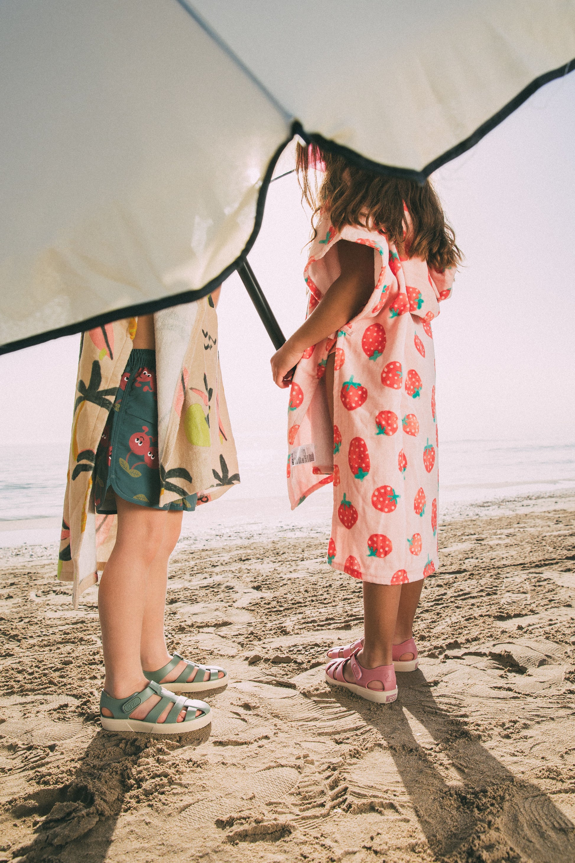 A child's closed toe unisex jelly sandal by Igor, style Bay, in nuevo rosa with cream sole with velcro fastening. Lifestyle image of chldren on beach.