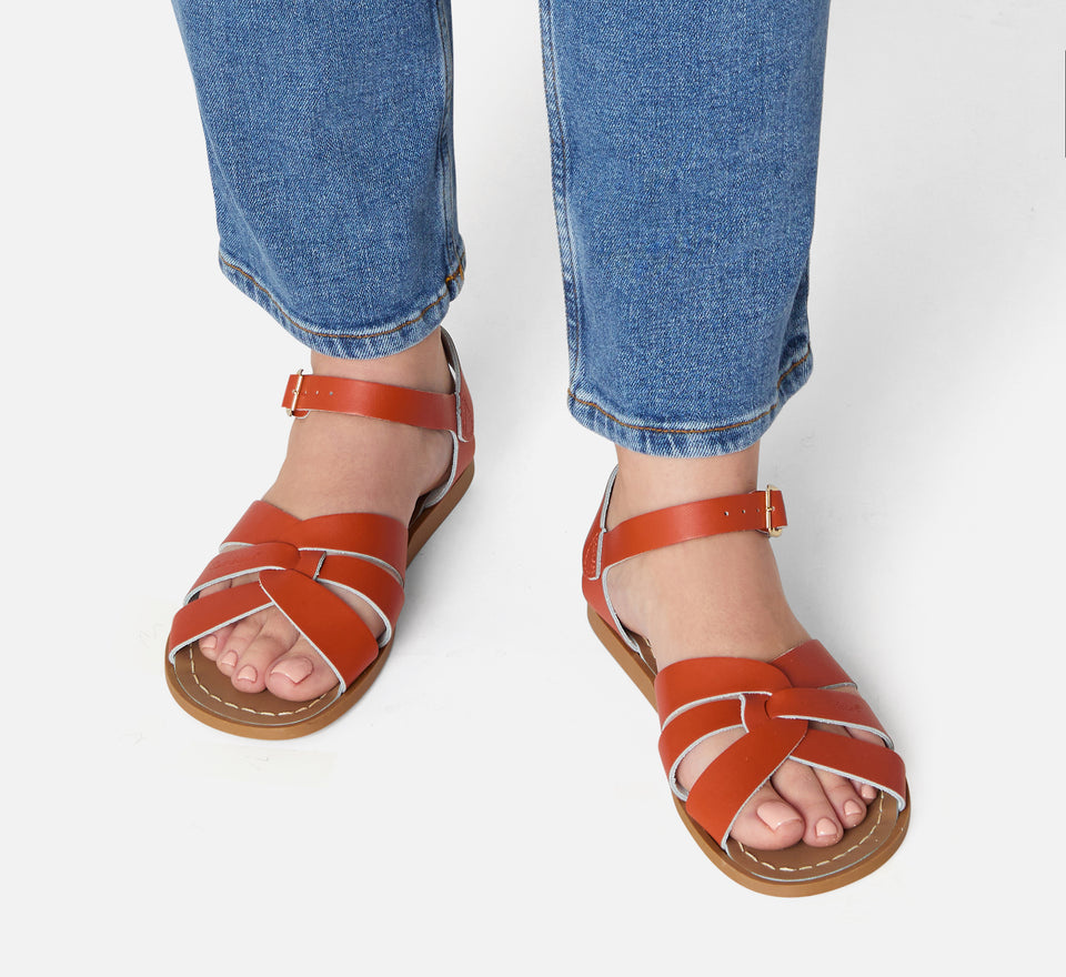 An original girls sandal by Salt Water Sandals in paprika with a single buckle fastening around the ankle. Open Toe and Sling-back with woven detail on the front. Lifestyle of looking down view.