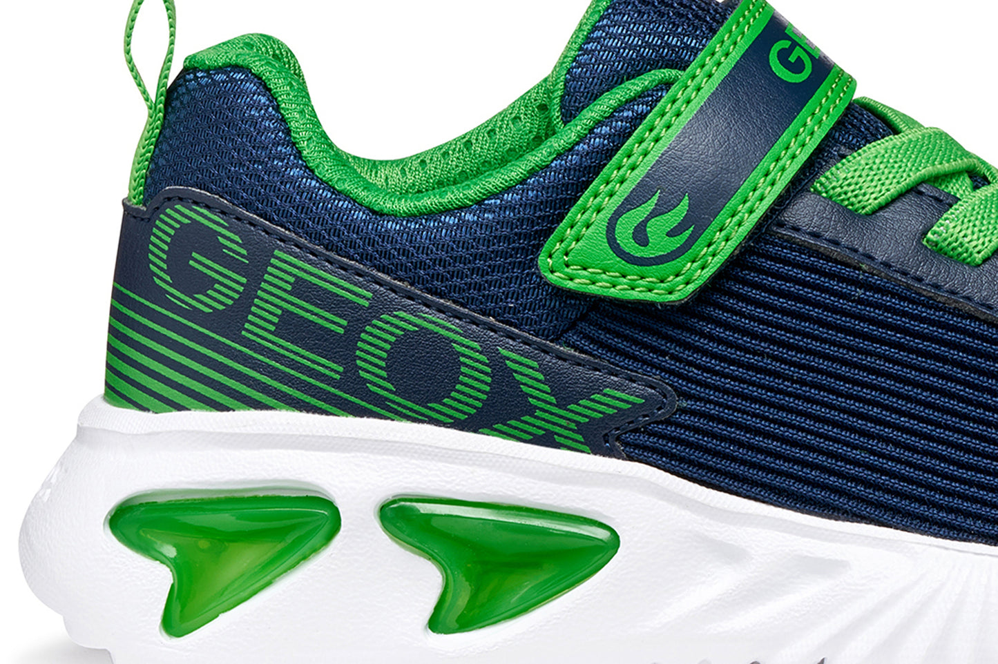 A boys trainer by Geox style Assister in navy/green with velcro strap, elastic laces, large geox logo on the side and lights in white sole. Zoomed image of Geox logo on side