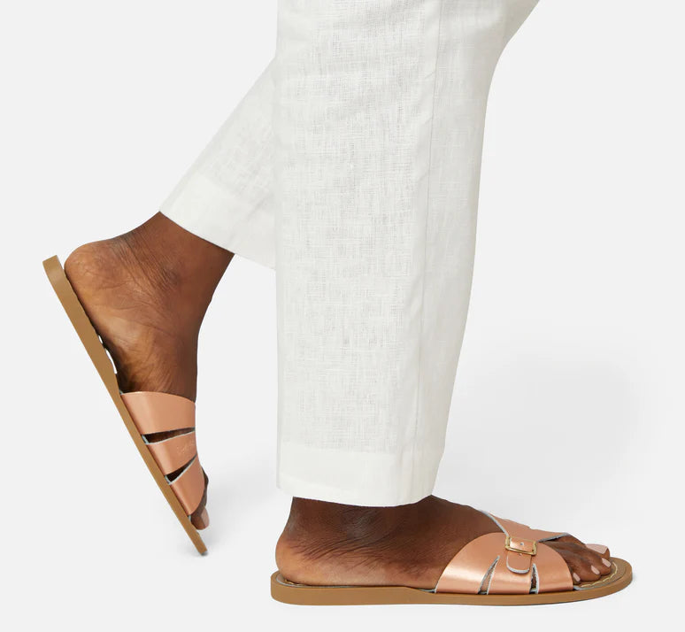 A Slide sandal by Salt Water Sandals in rose gold with single buckle fastening across the instep. Open toe and open back. Woman walking lifestyle view.