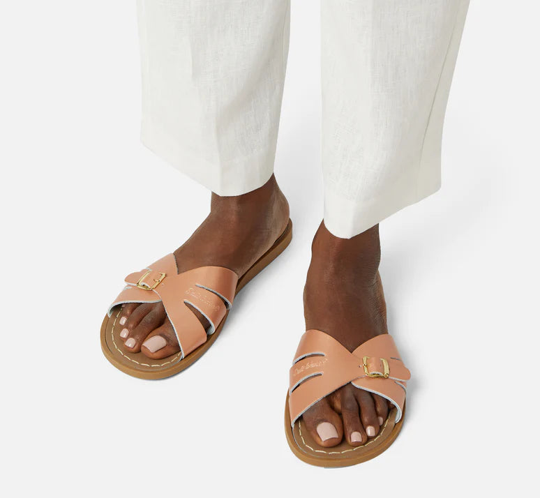 A Slide sandal by Salt Water Sandals in rose gold with single buckle fastening across the instep. Open toe and open back. Woman standing lifestyle view.