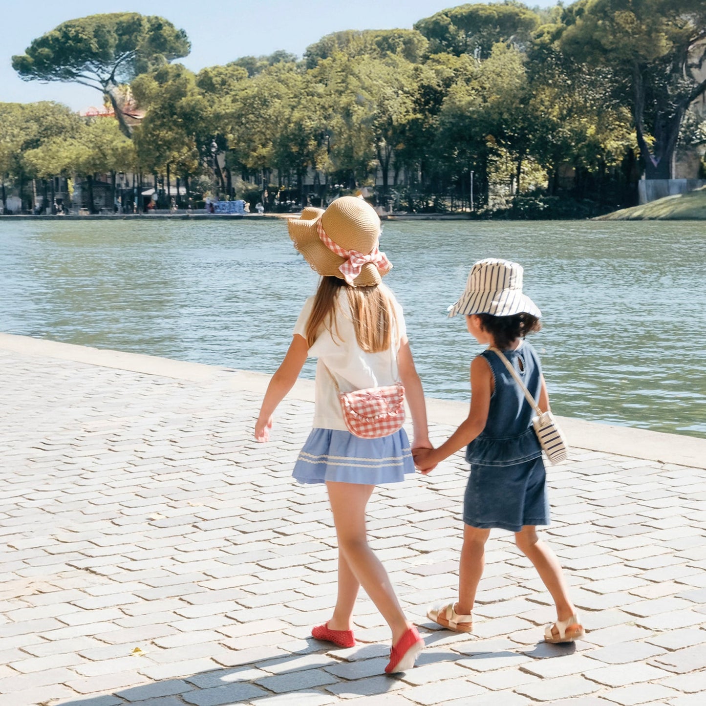 Two children holding hands by a lake, wearing Rockahula sun hats.