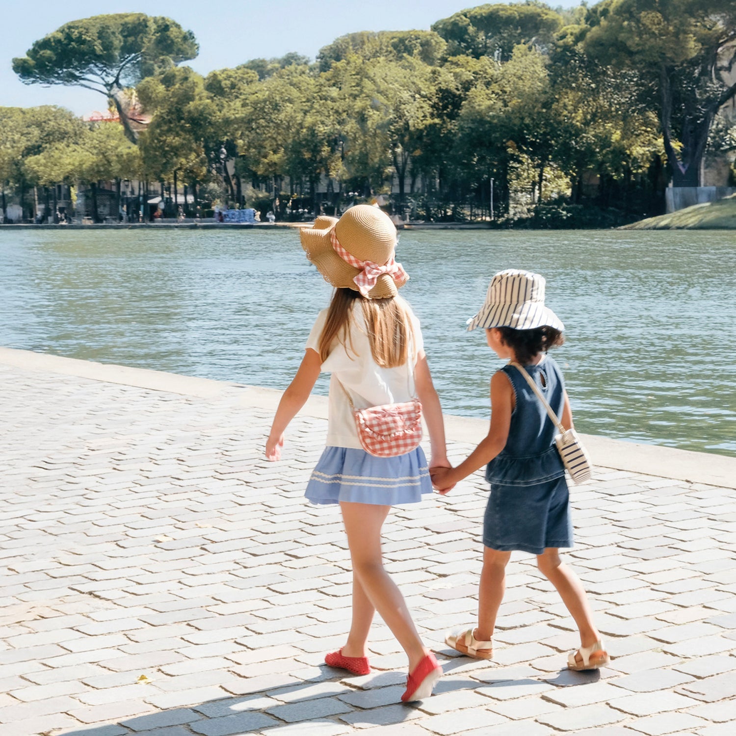 Two children holding hands by a lake, wearing Rockahula sun hats.