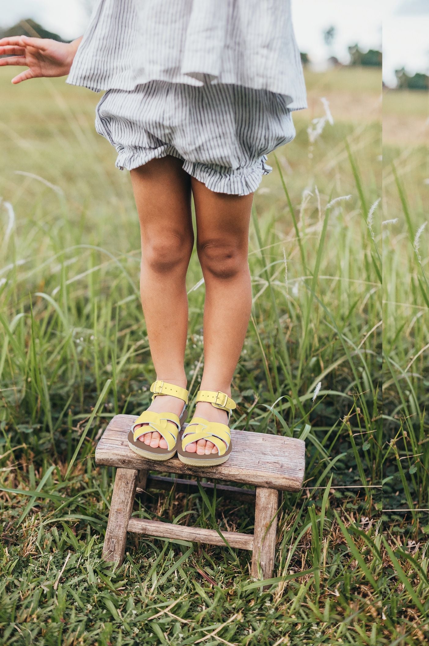 A girls sandal by Salt Water, style Swimmer in butter yellow, buckle fastening. Lifestyle of girls legs standing on a stool wearing sandals.