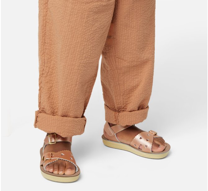 A girls sandal by Salt Water Sandal in rose gold with double buckle fastening across the instep and around the ankle. Featuring scallop edge and punched out heart detail. Lifestyle view.